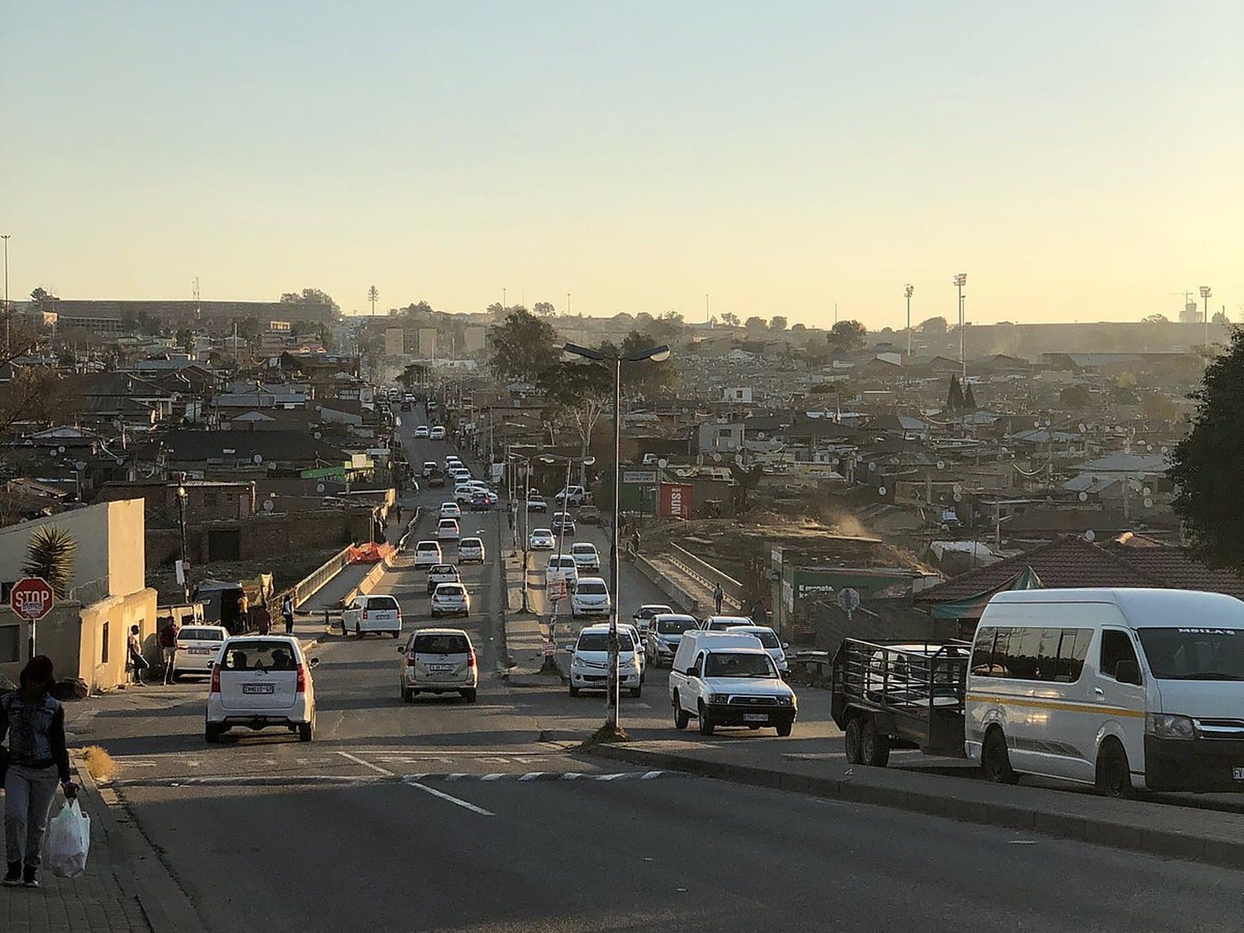 A main road through Alexandra in the evening light, traffic moving in both directions and a woman with a shopping bag on the pavement.
