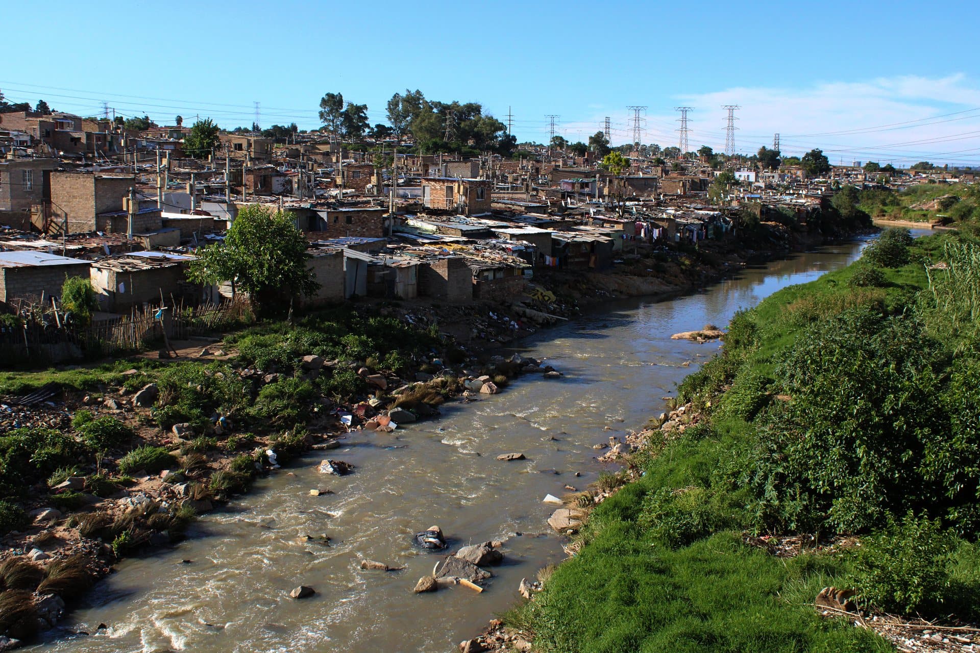 The Jukskei River winding past homes on the edge of Alexandra, power lines and a clear blue sky above.
