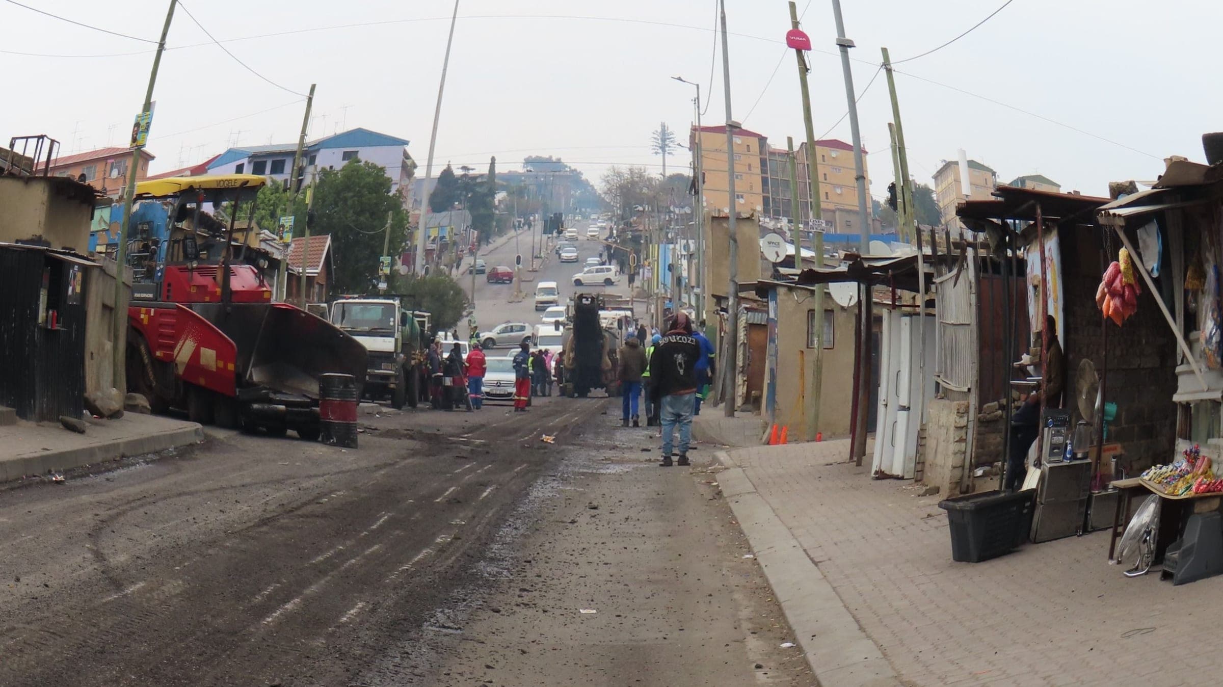 A crew resurfacing a steep Alexandra road, a paver and trucks at work while neighbours walk by and traders sit beside their stalls.