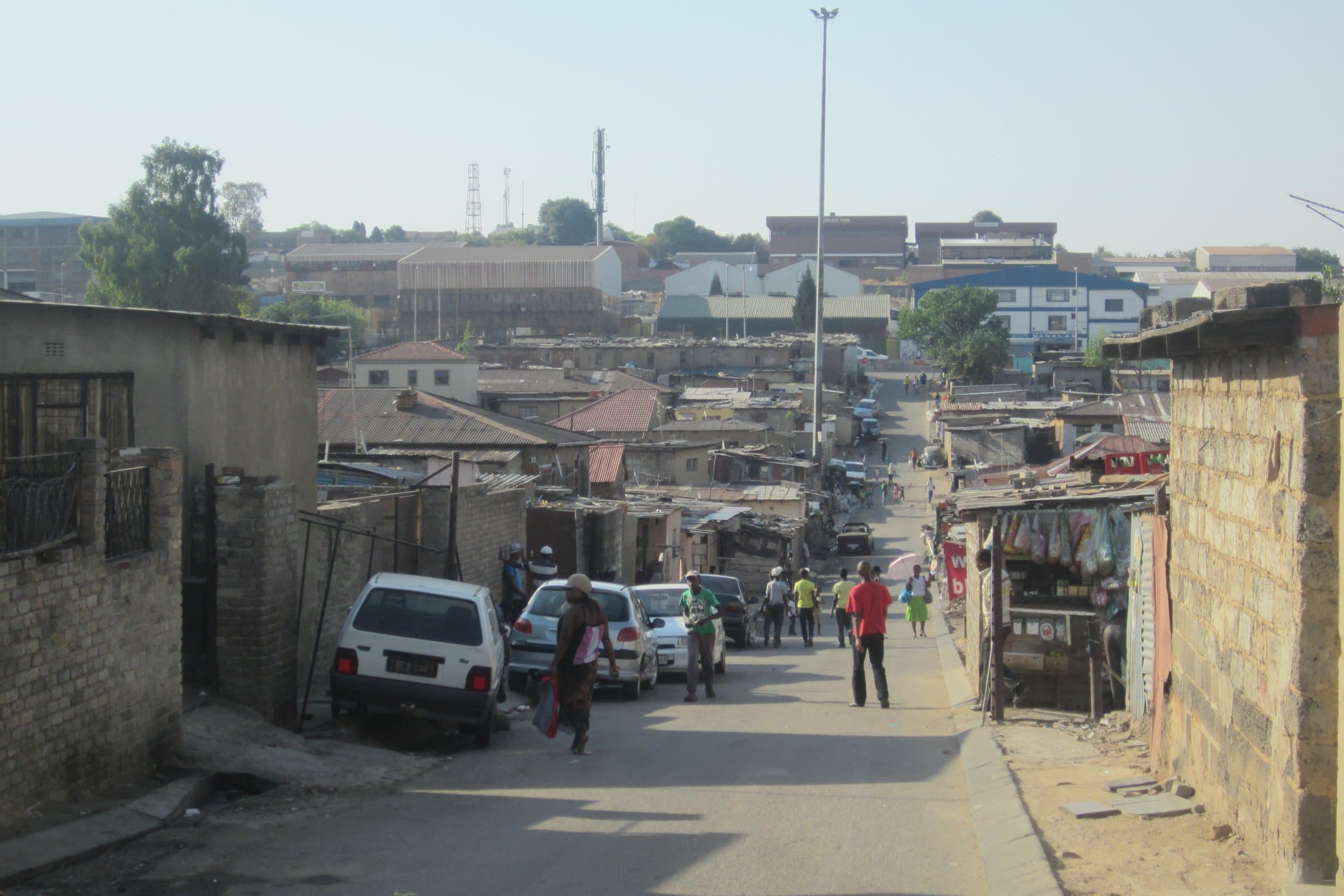 People walking up a busy Alexandra street in the afternoon sun, parked cars and a small shop on the right and the township's rooftops beyond.