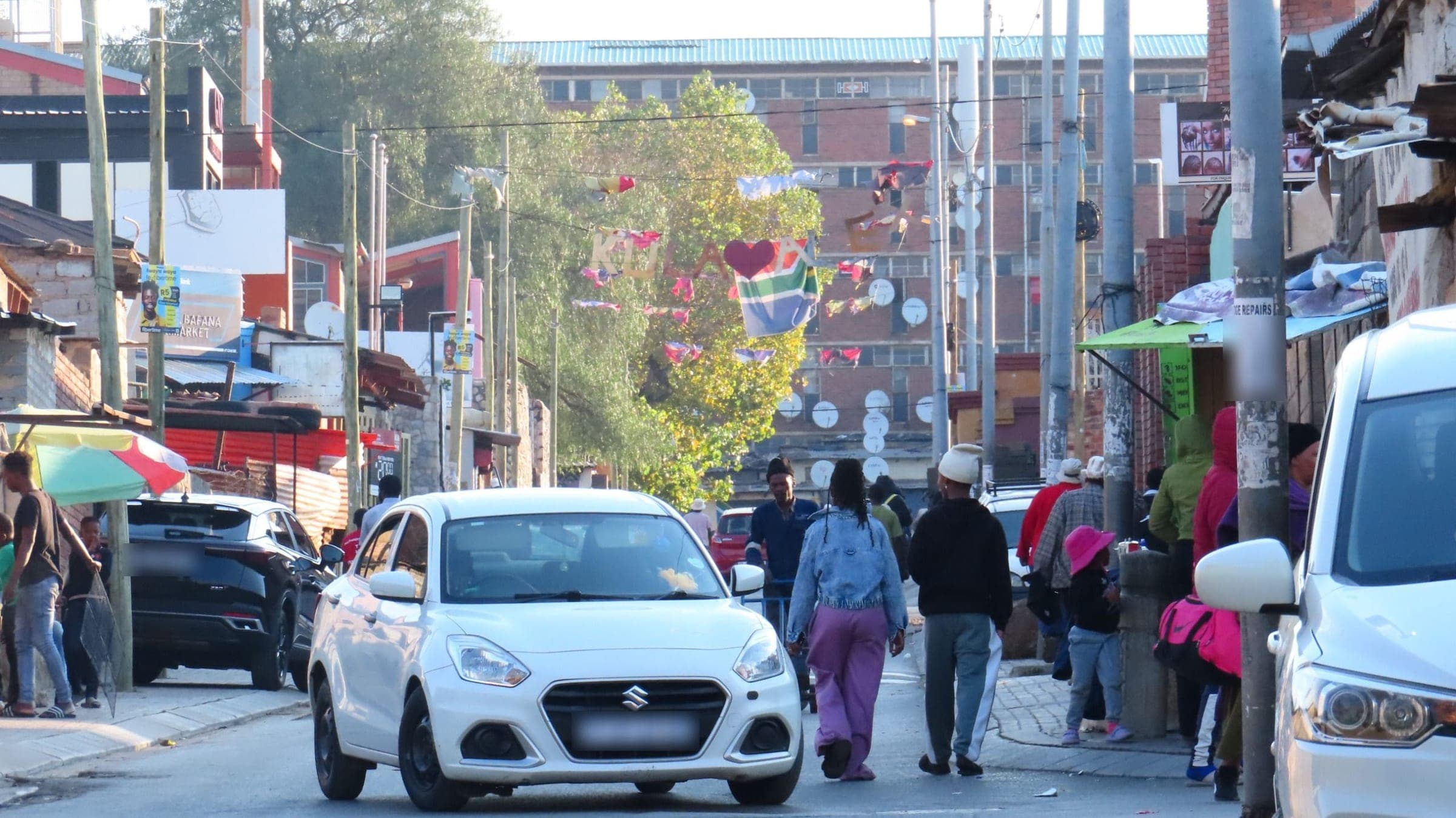 A busy Alexandra street in the evening light, neighbours walking past parked cars beneath strings of flags and bunting, a red-brick block rising behind the trees.