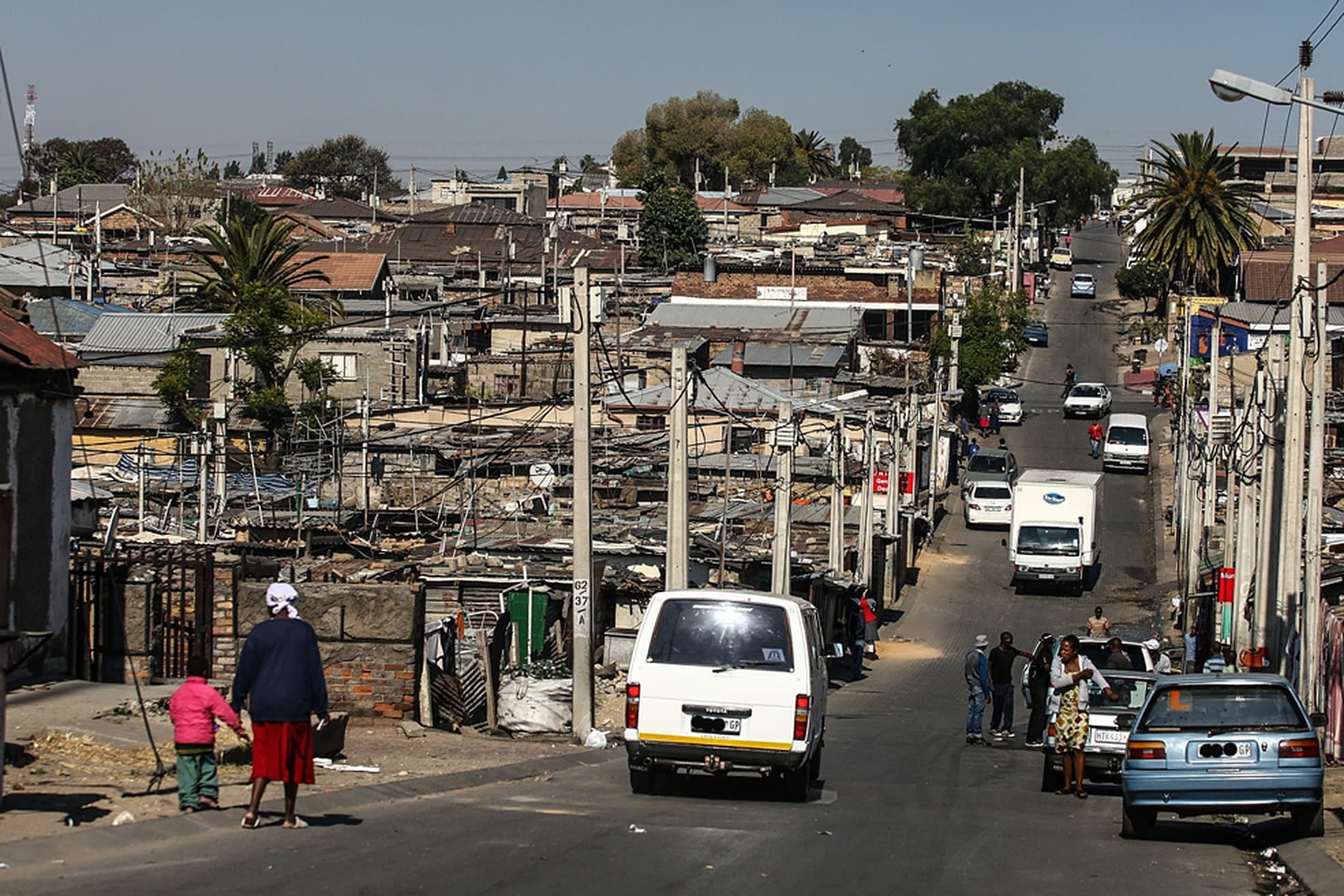 An Alexandra street climbing away from the camera, a woman and a child walking in the foreground, taxis and cars on the road and neighbours talking beside them.