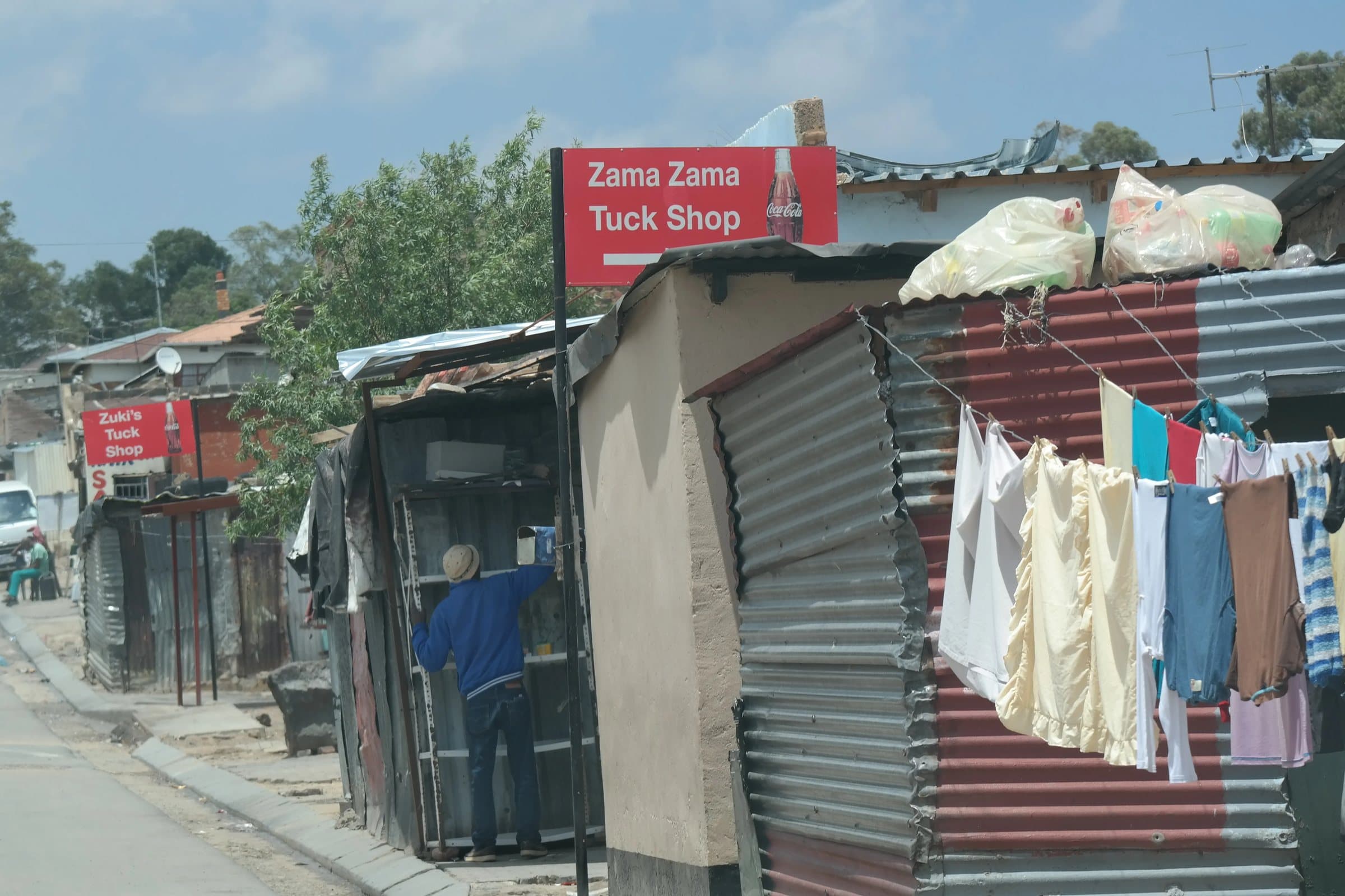A man restocking the shelves of a corrugated-iron tuck shop under a red sign, washing hanging on the line beside it.