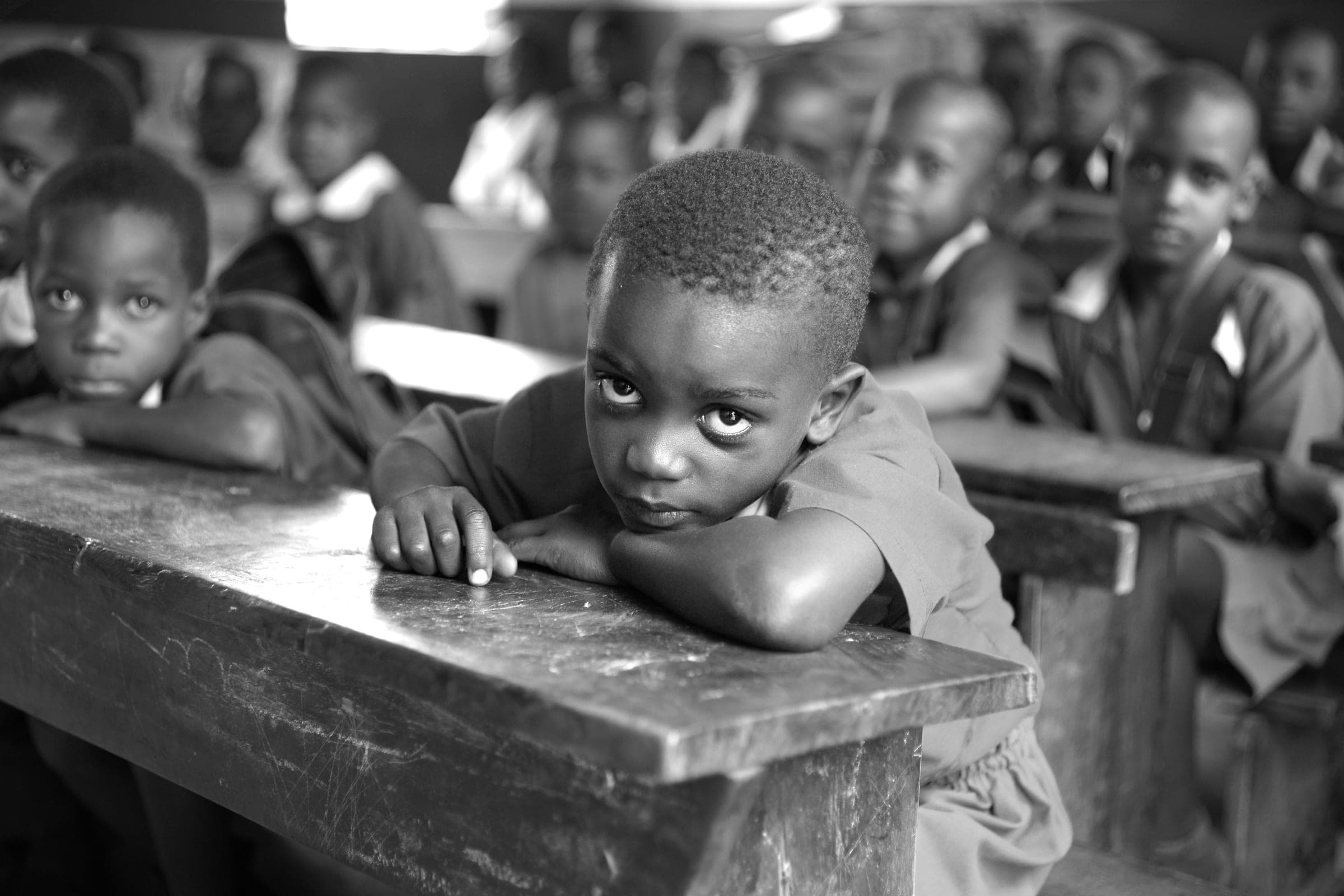 A boy resting his arms on his desk in a full classroom, looking straight ahead.