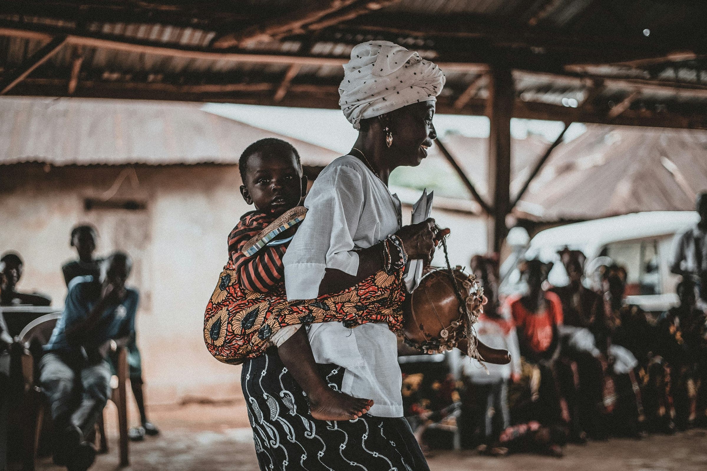A woman carrying her baby on her back and a basin on her head, walking through a covered market.