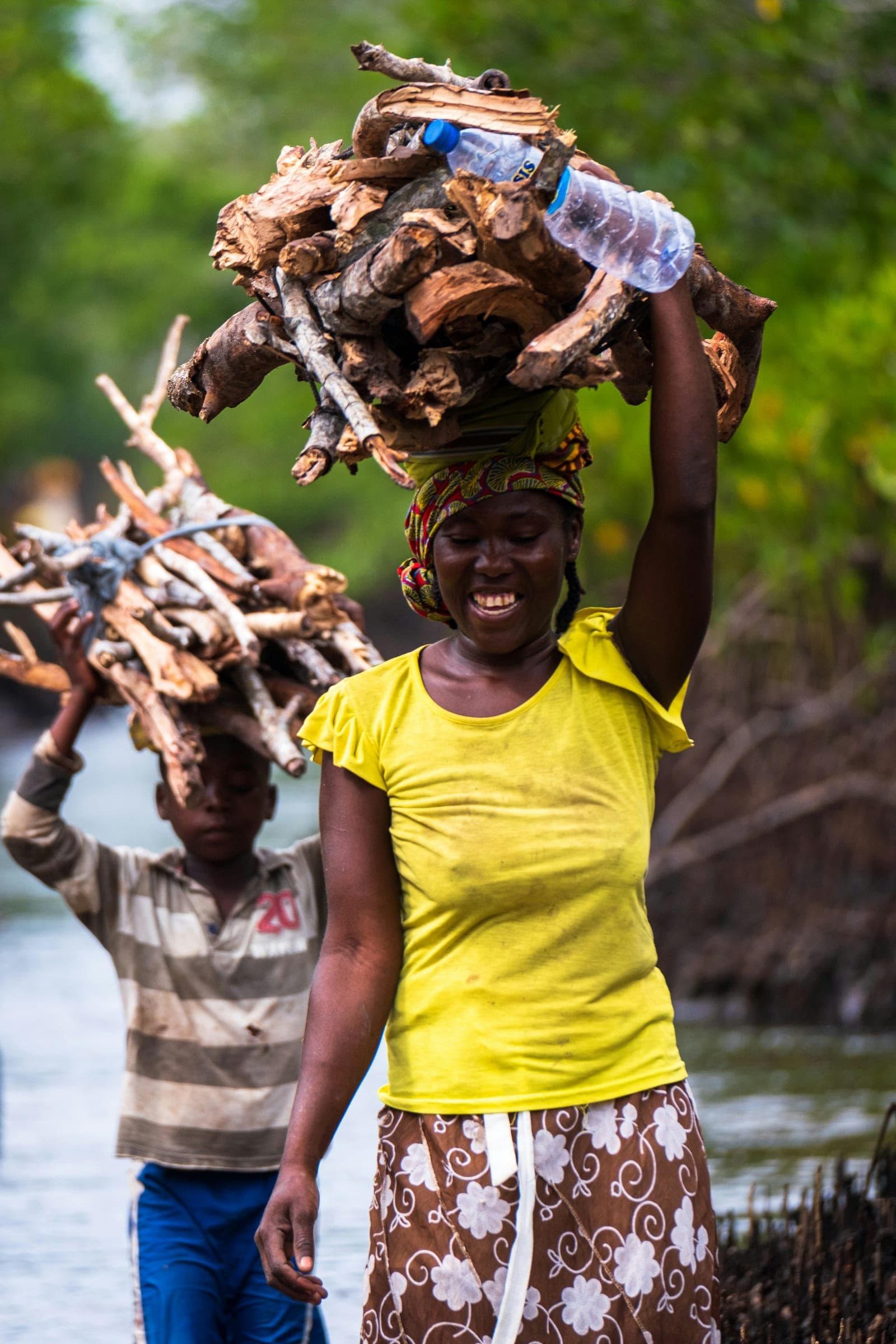 A woman carrying a bundle of firewood on her head, laughing, a child walking beside her.