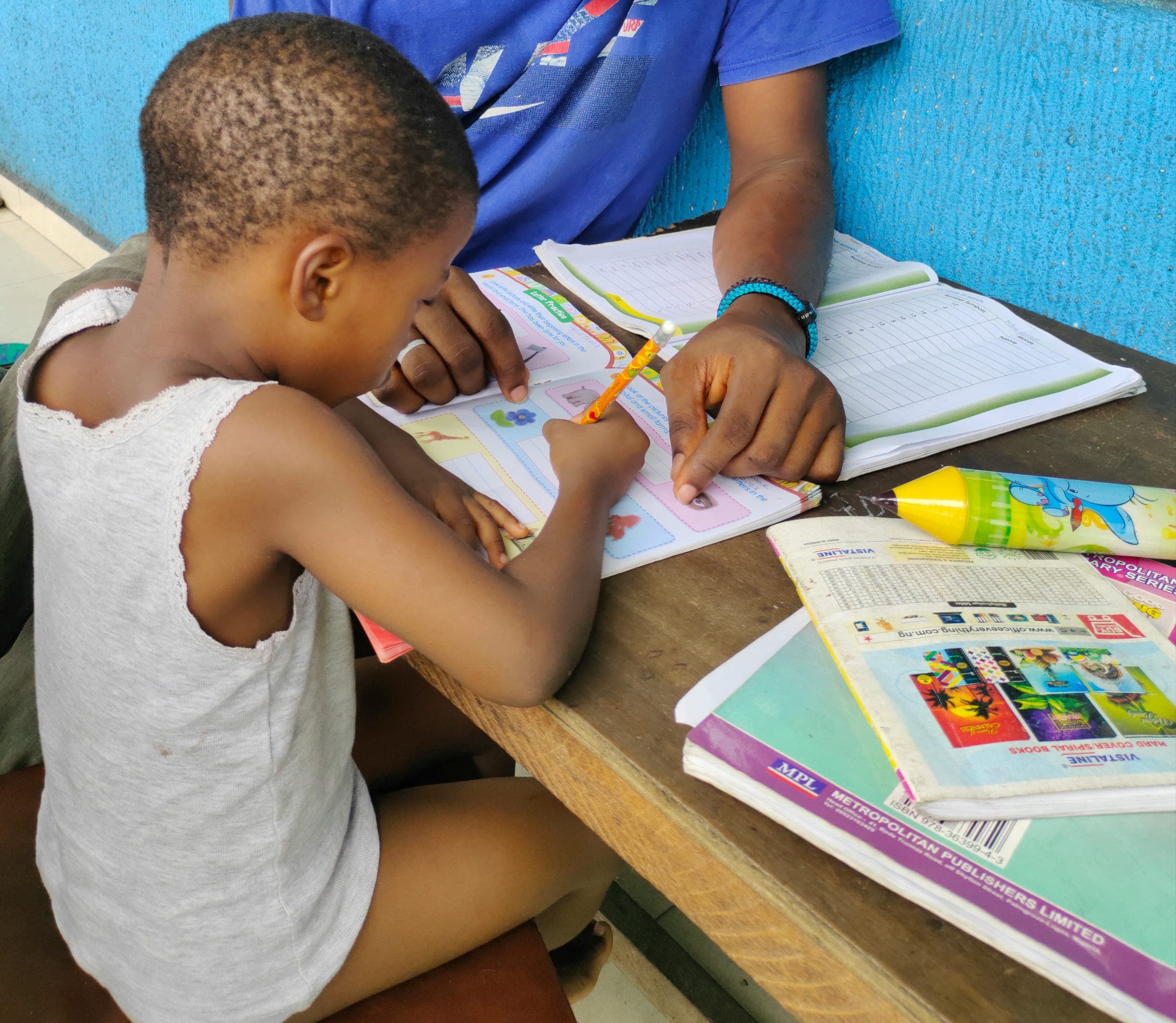 A child colouring carefully in a workbook at a desk, pencils and books spread out beside them.