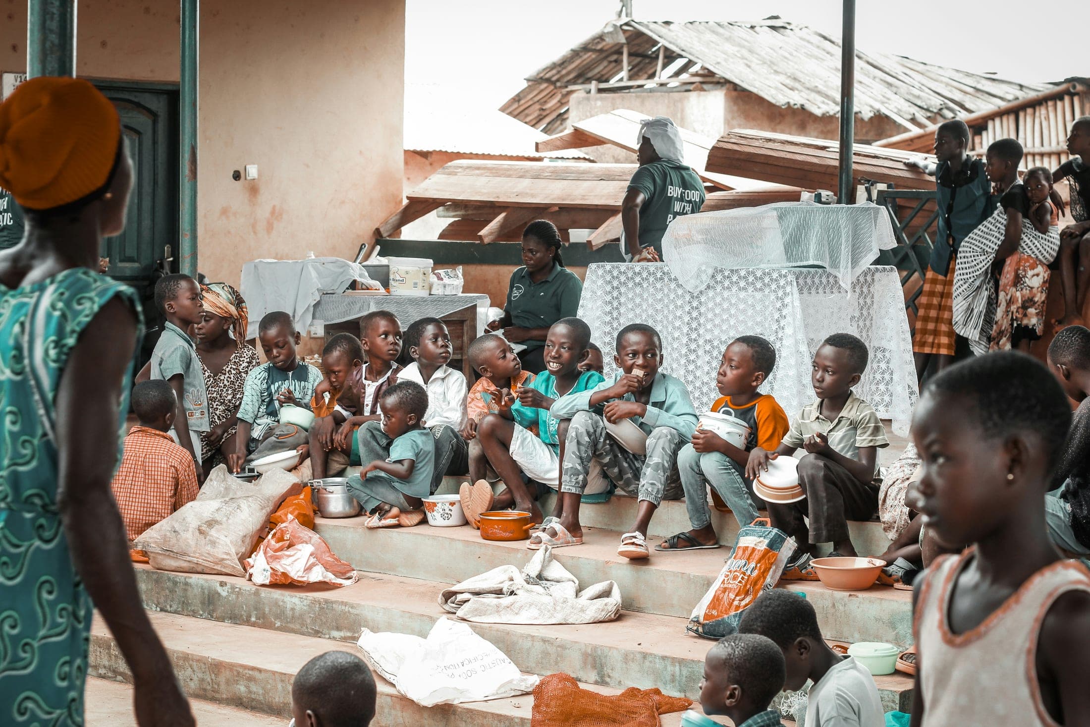 Children sitting together on steps with bowls, sharing a meal, while adults serve and watch from behind.