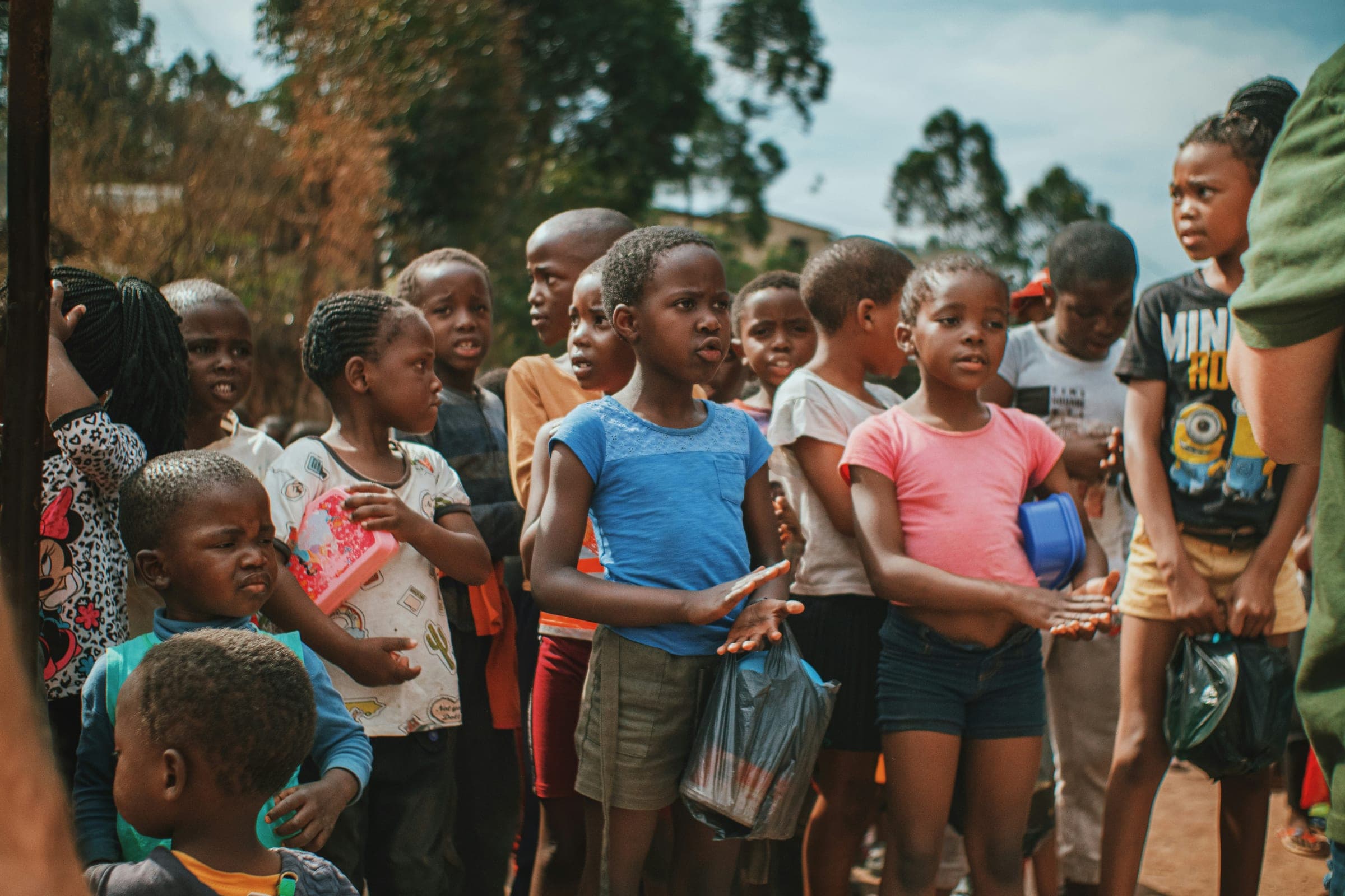 A crowd of children waiting together in a line outdoors, watching something out of frame.