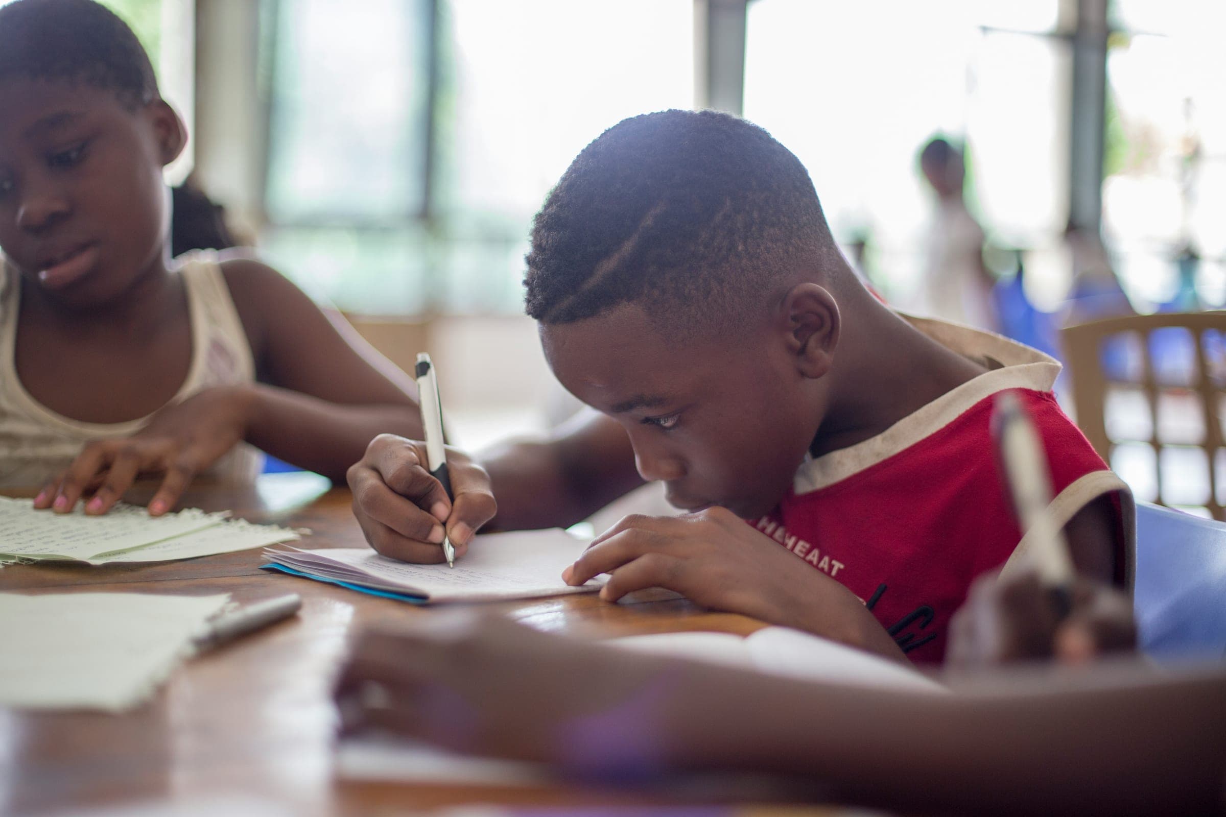 Two children working side by side at a table, writing in their books.