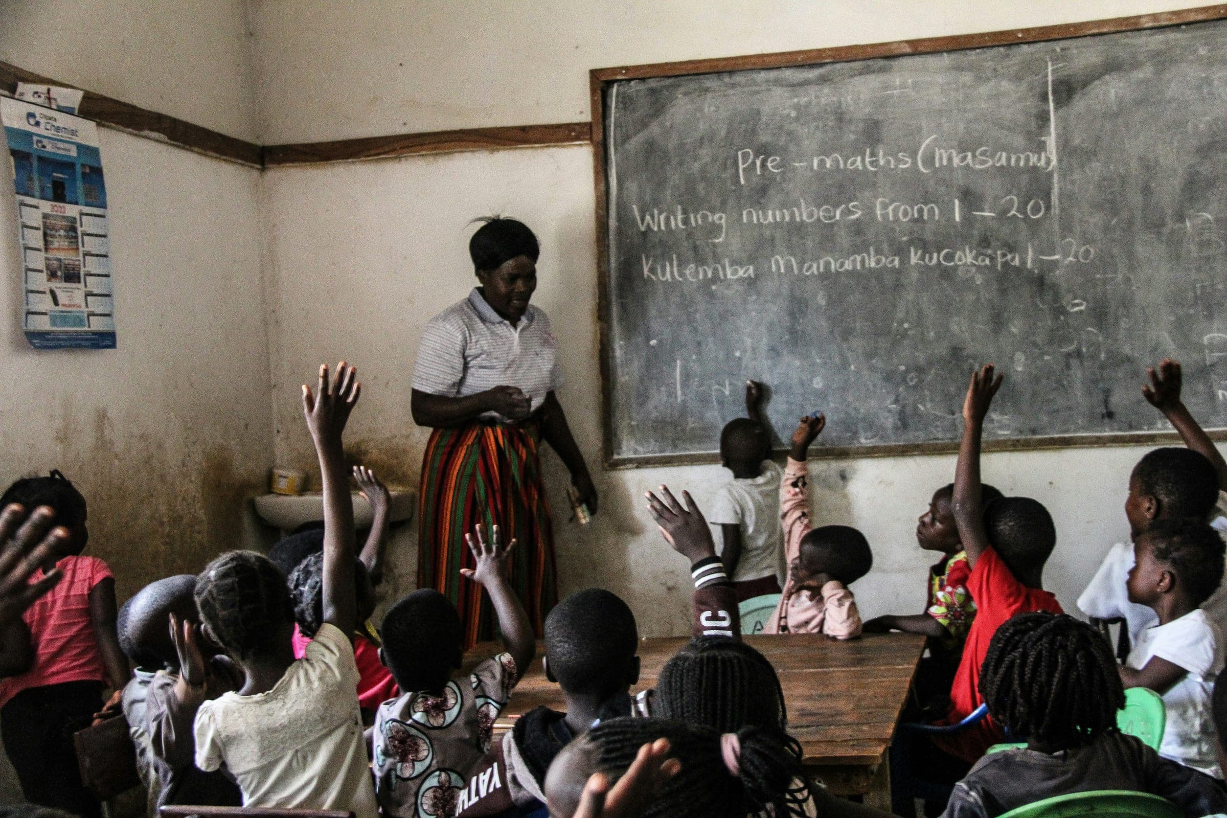 A teacher at the blackboard and a classroom of children with their hands up to answer.