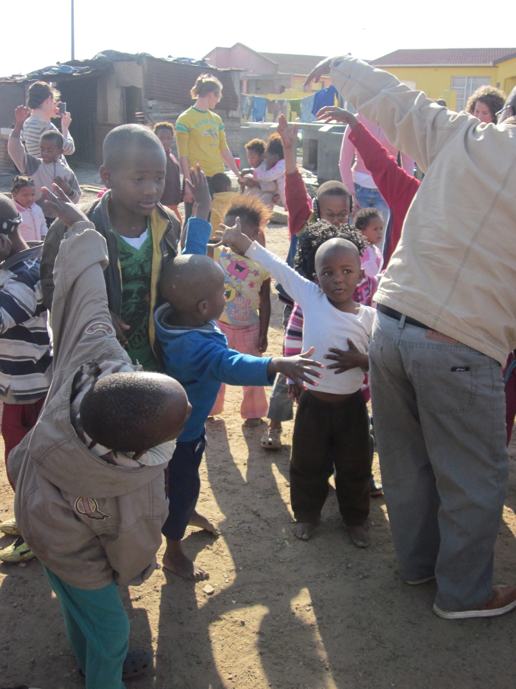 Children and adults together in an open yard, arms raised mid-game, homes rising on the slope behind them.