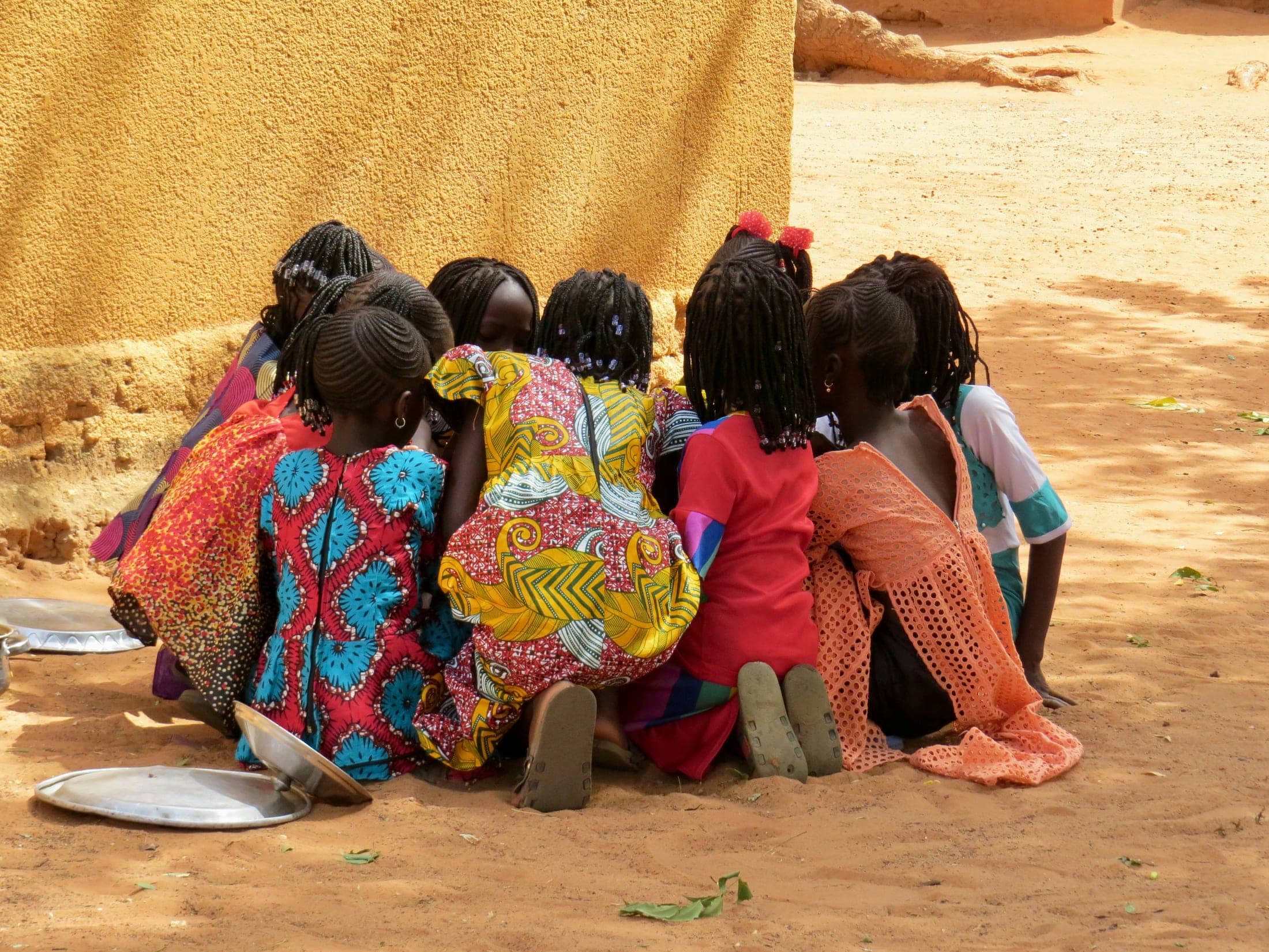 A group of girls crouched together in a circle in a sunlit courtyard, heads bent over something between them.