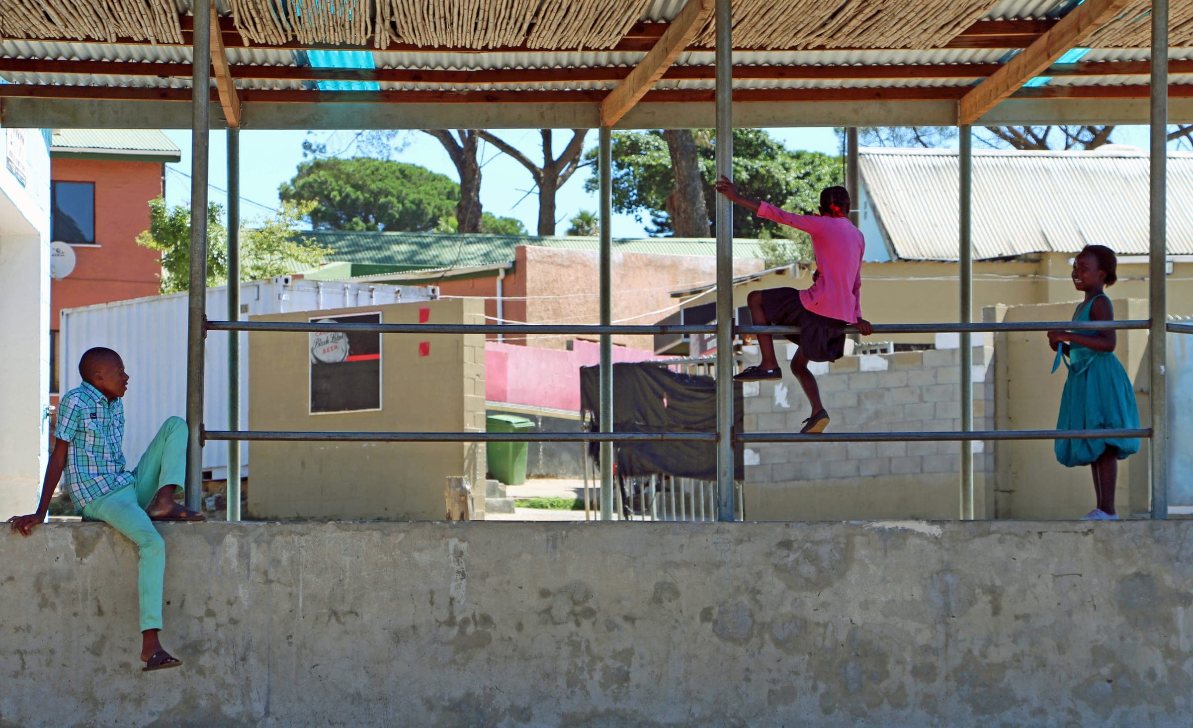 Children of Kayamandi playing under the shade structure of a community building, homes rising on the hillside behind them.