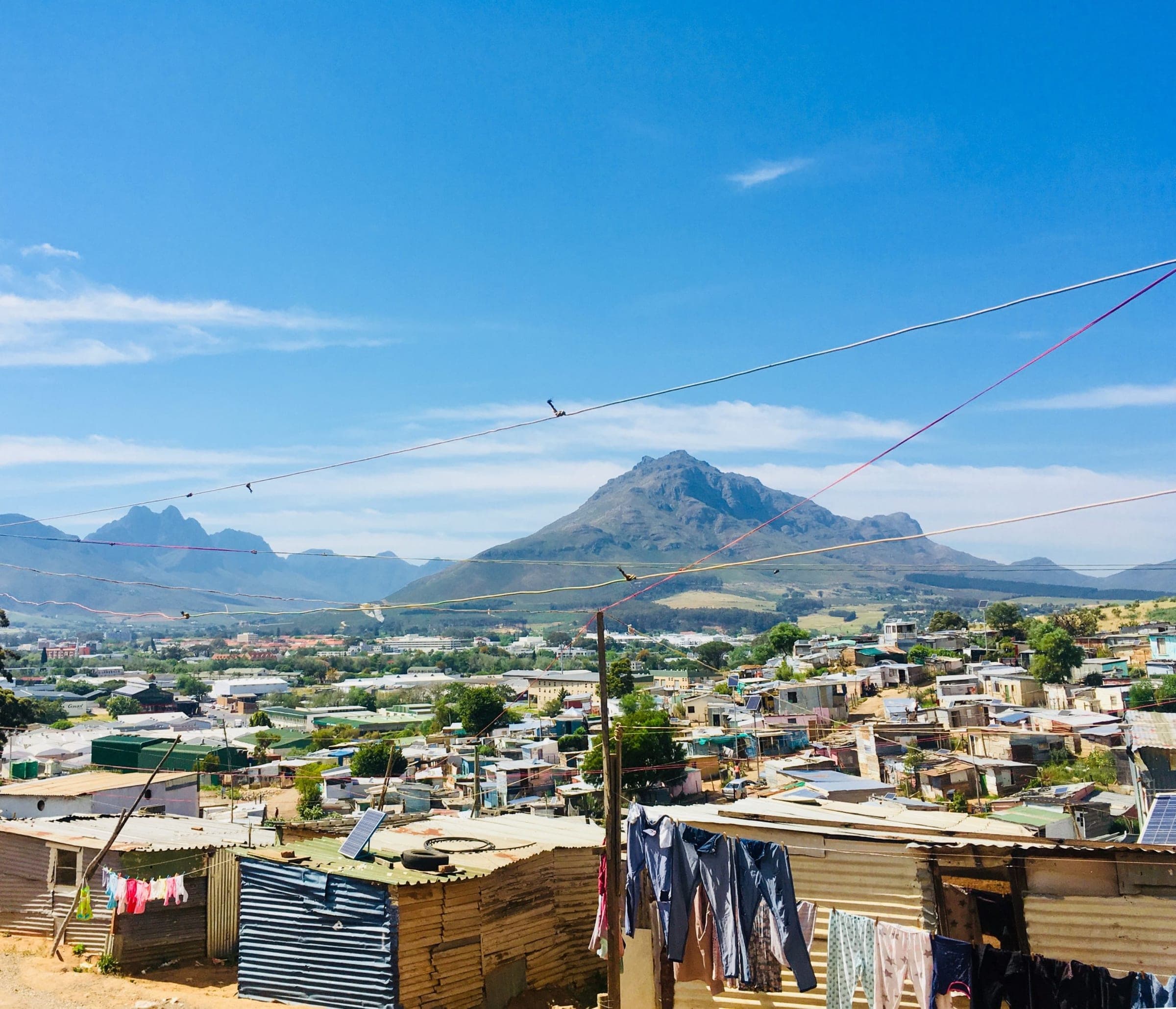 Kayamandi seen from the slope above it — rooftops running down toward Stellenbosch, washing out on the line in the foreground and the mountains beyond.