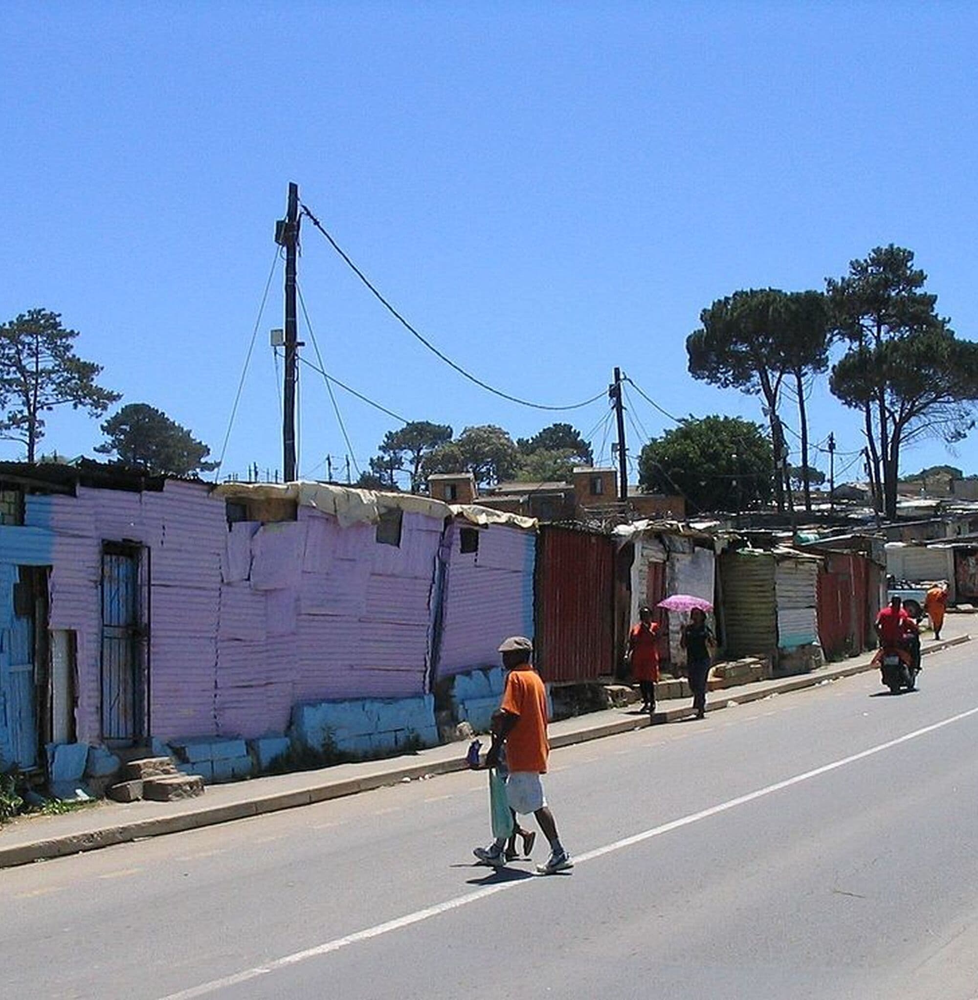 A Kayamandi street on a clear day, painted homes along one side and a resident walking in the road.