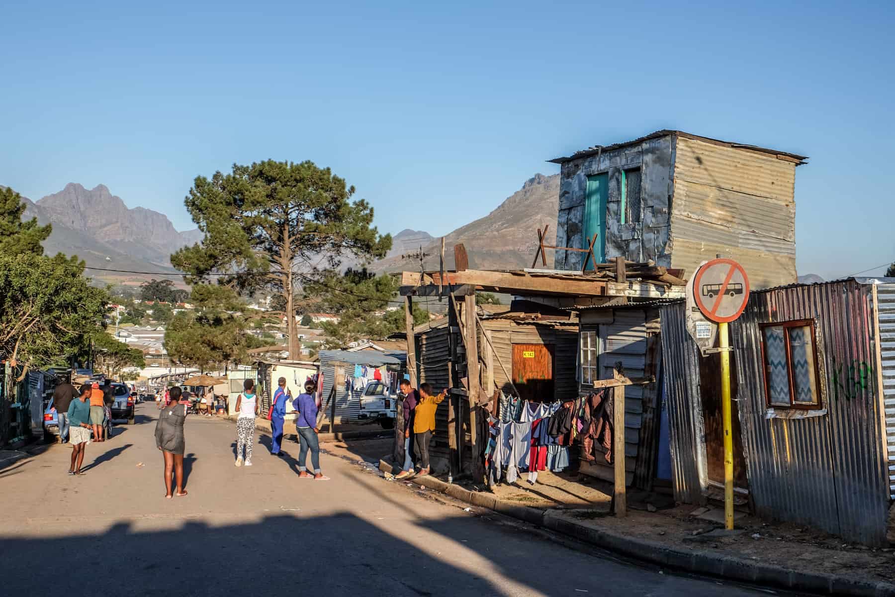 Neighbours walking a wide sand road between homes in Kayamandi, with the Boland mountains rising behind the rooftops.