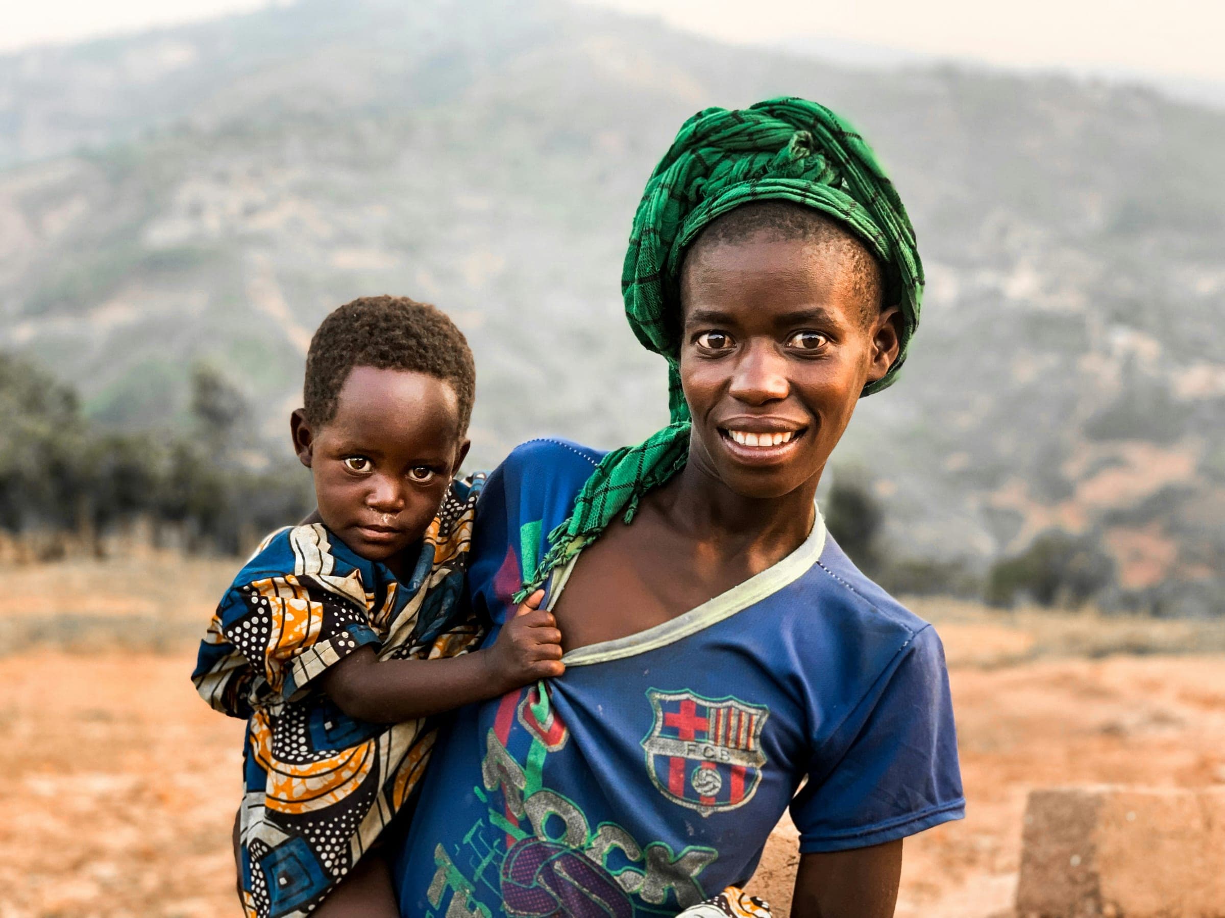 A young mother in a green headwrap carrying her small child, both looking toward the camera, the hills behind them.